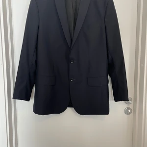 Svart kavaj från Filippa K, storlek 56 - Stilren svart kavaj från Filippa K i klassisk modell med två knappar framtill och fyra dekorativa knappar vid ärmslutet. Kavajen har två fickor med lock och en bröstficka. Materialet är slätt och känns lyxigt, perfekt för en snygg look.