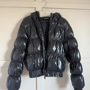 Svart glansig pufferjacka Brave Soul - Säljer en svart, glansig pufferjacka från Brave Soul. Jackan har en croppad passform, huva och dragkedja framtill. Den är vadderad och har elastiska muddar vid ärmslut och midja för extra komfort. Perfekt för kalla dagar när du vill sticka ut.