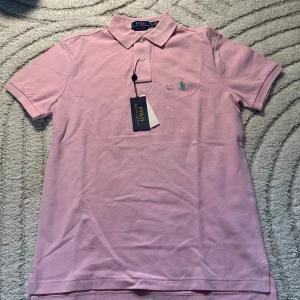 Rosa pikétröja från Polo Ralph Lauren - Säljer en ljusrosa pikétröja från Polo Ralph Lauren i bomull. Klassisk krage, två knappar och den ikoniska gröna loggan broderad på bröstet. Tröjan har korta ärmar och en slim fit-passform. Perfekt för en clean och stilren look.