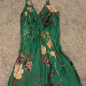 Grön blommig kortklänning från Zara - Supersöt grön kortklänning från Zara med blommigt mönster i beige, lila och brunt. Klänningen har smala axelband, v-ringning både fram och bakoch rynkdetalj framtill. Använd ett fåtal gånger och är i bra skick. Storlek xs men skulle säga att den även passar s. 100% viskos.