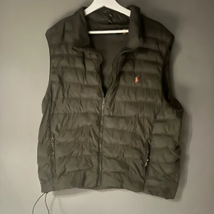 Olivgrön dunväst från Polo Ralph Lauren - Snygg olivgrön dunväst från Polo Ralph Lauren med klassisk quiltad design och dragkedja framtill. Västen har två fickor med dragkedja och ikoniska broderade loggan på bröstet. Perfekt att slänga över en hoodie eller tröja för extra värme.  dragkedjan till höger ytterficka är trasig men går troligen att laga utan problem.