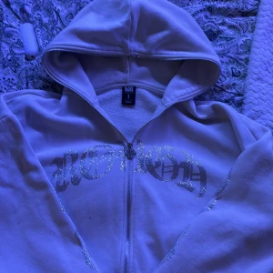 Vit hoodie med strass och flames - Vit hoodie från Black Squad i oversized fit med glittriga strassdetaljer. Text i silver på bröstet och flames längs båda ärmarna. Dragkedja framtill med unik zip puller och stor huva. Perfekt statement-plagg för dig som gillar streetstyle.