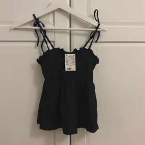 Svart smockad linnetopp med knytband - Supersöt svart linnetopp från H&M med smockad överdel och volangkant upptill. Toppen har tunna axelband som knyts upptill för en justerbar passform. Perfekt till sommaren och enkel att styla med jeans eller kjol.