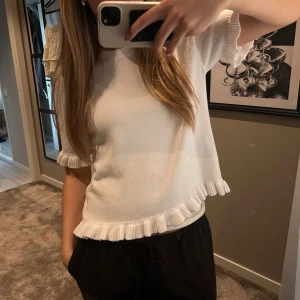 Vit stickad topp med volangkant - Supersöt vit stickad topp med korta ärmar och volangdetaljer längs ärmslut och nederkant. Modellen är croppad och har en avslappnad passform som känns både trendig och bekväm. Perfekt att styla med jeans eller kjol för en fräsch look.