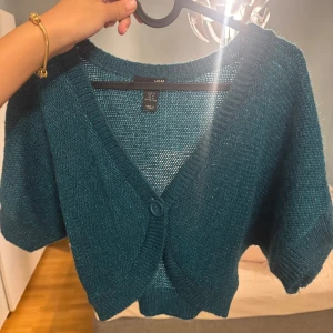 Bolero  - Säljer en turkos stickad bolerotopp från H&M med korta ärmar och v-ringning. Toppen har en stor knapp framtill och är croppad i modellen. Perfekt att slänga över ett linne eller klänning för en cool lager-på-lager-look.