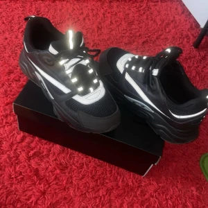 Svarta b22 sneakers med reflexdetaljer - Säljer ett par B22 sneakers i svart med vita och reflekterande detaljer. Skorna har en kraftig sula, mesh och skinnpaneler samt snörning framtill. Perfekta för dig som vill sticka ut med exklusiva och trendiga sneakers.