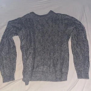 Grå stickad kabeltröja - Mysig grå stickad tröja med klassiskt kabelstickat mönster. Tröjan har rund halsringning och långa ärmar med ribbade muddar. Perfekt för kyliga dagar och enkel att matcha med jeans eller kjol.