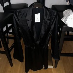 Svart satin klänning DW Freedom - Snygg svart klänning i satin från DW Freedom. Klänningen har lång ärm, markerad midja och klassisk krage med v-ringning. Perfekt för dig som vill ha en stilren och elegant look. Finns i storlek M.