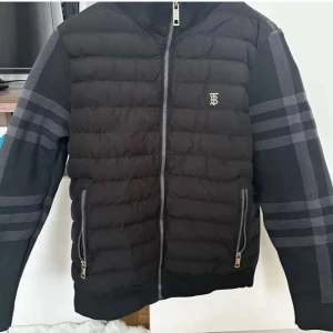 Svart pufferjacka från Burberry - Snygg svart pufferjacka från Burberry med klassiskt rutmönster på ärmar och rygg. Jackan har quiltad framsida, dragkedja framtill och två fickor med dragkedja. Materialmix med polyester och typisk Burberry-detalj på bröstet. Perfekt för dig som vill ha en stilren och exklusiv look.