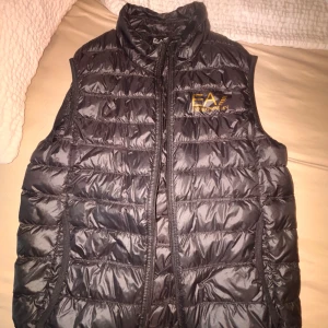 Svart väst EA7 Emporio Armani - svart väst från EA7 Emporio Armani med guldfärgad logga på bröstet. Västen är quiltad och har dragkedja framtill. Perfekt att slänga över en hoodie eller tröja på sig.
