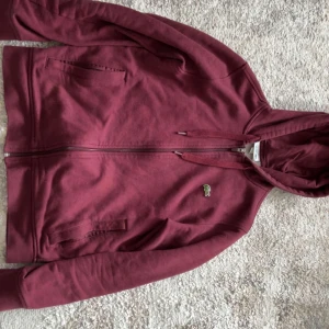 Vinröd Lacoste Hoodie - Säljer en vinröd hoodie från Lacoste Sport med dragkedja och huva. Tröjan har klassisk krokodillogga på bröstet, två fickor framtill och justerbara snören i huvan. Perfekt för chill dagar eller när du vill ha en sportig look.