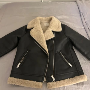 Svart teddyjacka från Mango med fuskpäls - Säljer en svart teddyjacka från MNG med mjukt beige fuskpälsfoder och krage. Jackan har två snedställda fickor med dragkedja, silverfärgade detaljer och breda muddar i fuskpäls. Perfekt för kalla dagar och riktigt snygg till jeans eller kjol.