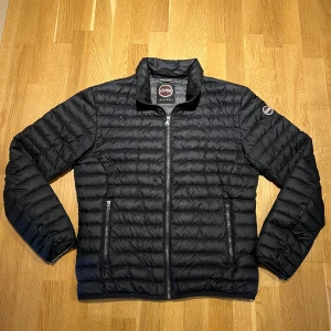 Svart dunjacka från Colmar - Snygg svart dunjacka från Colmar med horisontella sömmar och diskret logga på ärmen. Jackan har dragkedja framtill och två dragkedjeförsedda fickor. Perfekt för kyliga dagar och har en stilren, sportig look.