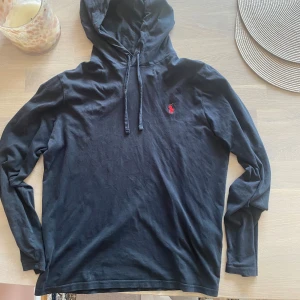 Svart hoodie från Polo Ralph Lauren - Svart hoodie från Polo Ralph Lauren i mjuk bomull med klassisk huva och snörning. Tröjan har lång ärm och den ikoniska röda loggan broderad på bröstet. Perfekt för en chill och stilren look.