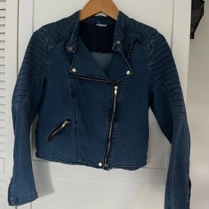 Mörkblå jeansjacka med dragkedjor - Snygg mörkblå jeansjacka i bikerstil med markerade axlar och ärmar, guldfärgade dragkedjor och detaljer. Jackan har croppad passform och coola sömmar på axlarna för extra edge.