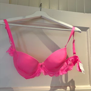 Chockrosa spets-bh med vaddering - Säljer en chockrosa bh med vadderade kupor och justerbara axelband. Bh:n har fina spetsdetaljer längs kanterna och sidorna, aldrig andvänd. 