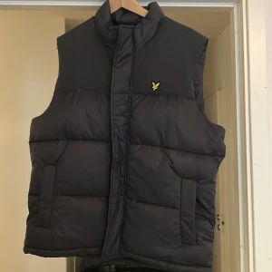 Svart dunväst från Lyle & Scott, strl L - Svart dunväst från Lyle & Scott med gul broderad logga på bröstet. Västen har hög krage, två sidofickor och quiltad design. Materialet är polyester och västen har en stilren, normal passform. Perfekt för lager-på-lager under kyliga dagar.