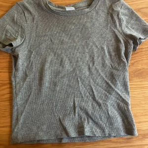 Grå ribbad t-shirt från Lindex - Grå ribbad t-shirt från Lindex i storlek 158/164. T-shirten har korta ärmar och en rund halsringning. Materialet är mjukt och stretchigt, perfekt för en chill och enkel look. Passformen är normal och färgen är neutral grå.