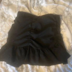 Svart volangkjol med rynkade detaljer - Säljer en svart kortkjol med volangkant och rynkade detaljer framtill. Kjolen har en feminin och trendig look, perfekt att styla med en crop top eller stickad tröja. Materialet känns mjukt och följsamt.