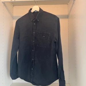 Svart långärmad skjorta i bomull - En svart skjorta från Filippa K i mjuk bomull med klassisk krage, knappstängning och en bröstficka. Skjortan har lång ärm och en clean, stilren look som funkar till många outfits. Perfekt för dig som gillar enkel och tidlös stil.