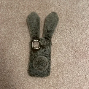iPhone 11 Fodral Kaninöron Päls - Sött och mjukt fodral till iPhone 11 med grå päls, kaninöron och svans. Kameraramen är dekorerad med strass och en liten rosett. Fodralet är i gott skick, perfekt för dig som vill ha ett unikt och lekfullt mobilskydd.
