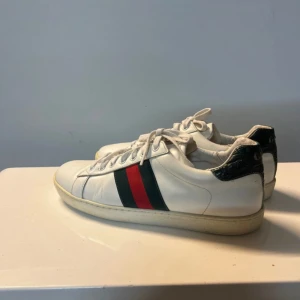 Gucci Ace vita sneakers med röd/grön rand - Gucci Ace sneakers i vitt skinn med klassiska gröna och röda ränder på sidorna. Baksidan har mörkgrön detalj med Gucci-logga och präglad struktur. Platt sula med unikt mönster undertill. Snygga och ikoniska sneakers som lyfter varje outfit.