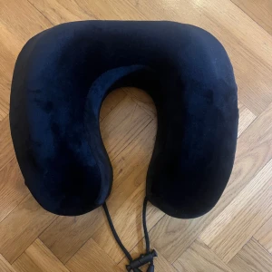 Nackkudde - Detta är en svart nackkudde i U-form, perfekt för resor och extra komfort. Kudden har ett justerbart snöre för att hålla den på plats. Tyget ser mjukt ut och kudden är i gott skick utan synliga defekter eller slitage.