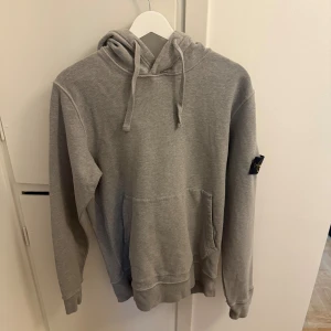 Grå hoodie från Stone Island - Säljer en grå hoodie från Stone Island med klassisk patch på vänster ärm. Tröjan har huva med snörning, magficka och ribbade muddar. Perfekt för dig som gillar streetwear och vill ha en stilren look.