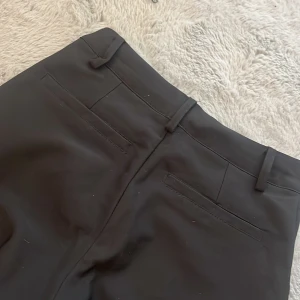 Svarta kostymbyxor från Xoxo Friends - Svarta kostymbyxor från Xoxo Friends med klassisk passform, bälteshällor och två bakfickor. Byxorna har knapp och dragkedja framtill och är tillverkade i ett slätt tyg som ger en stilren look. Perfekta för en dressad outfit.