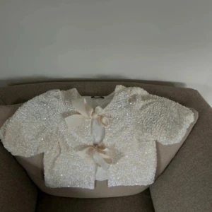 Glittrig topp med rosetter - Superfin glittrig topp i vit med två stora rosetter framtill. Koftan har kortare modell och puffiga ärmar, perfekt för att sticka ut. Materialet är täckt av paljetter som ger ett lyxigt intryck. Man kan även ha den bak&fram, super fint det också. 