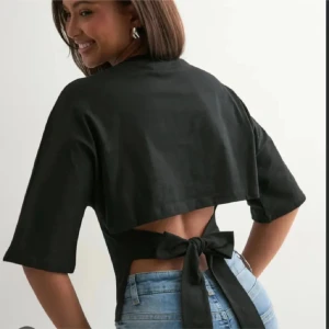 Svart croppad topp med knyt i ryggen Nelly - Säljer en svart croppad topp med korta ärmar och snygg öppen rygg som knyts med en stor rosett. Toppen har en loose passform och är perfekt för dig som vill ha något trendigt och bekvämt. Materialet känns som bomull eller linne. Från Nelly 