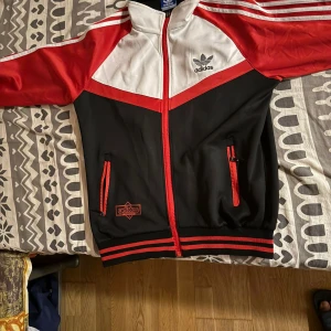 Adidas svart, röd och vit track jacket - Snygg Adidas track jacket med svart bas, röda ärmar och vita detaljer. Jackan har klassiska Adidas-ränder på axlarna, dragkedja framtill och två fickor med röda dragkedjor. Stor Adidas-logga på bröstet och en stor tryckt logga på ryggen. Perfekt för en sportig streetstyle.