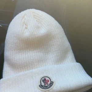 Vit ribbad mössa från Moncler - Snygg vit mössa från Moncler med ribbad struktur och uppvikt kant. Märkets klassiska logga sitter framtill. Mössan är stickad och har en clean, modern look som passar perfekt till kalla dagar.