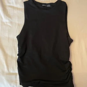 Svart ärmlös topp från SHEIN - Svart ärmlös topp från SHEIN med rund halsringning och rynkade detaljer längs ena sidan. Toppens enkla design gör den lätt att matcha och den har en tight passform som framhäver siluetten. Perfekt för dig som gillar stilrena basplagg.