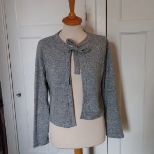 Grå kofta med knyt från 157 - Mysig grå kofta från 157 med knytband vid halsen. Koftan är i mjukt material, har lång ärm och en rak, lite croppad passform. Perfekt att slänga över en topp för en chill look.