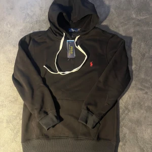 Svart hoodie från Polo Ralph Lauren - Svart hoodie från Polo Ralph Lauren med vit snörning och klassisk röd logga broderad på bröstet. Tröjan är i helt ny skick och prislappen finns fortfarande på. Storlek M. Skriv om du har några frågor.