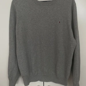 Grå stickad tröja Tommy Hilfiger - Klassisk grå stickad tröja från Tommy Hilfiger med rund halsringning och diskret logga broderad på bröstet. Tröjan har ribbade muddar vid ärmslut och nederkant, perfekt för lager-på-lager-stil. Enkel och stilren design som funkar till allt.