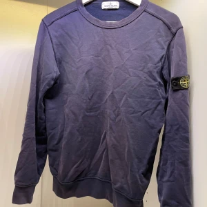 Mörkblå Stone Island tröja xs - Mörkblå tröja från Stone Island med den  klassiska loggan på ärmen. Tröjan har rund halsringning och långa ärmar. Tillverkad i mjuk bomull och har en avslappnad passform. Perfekt för dig som gillar streetwear och vill ha en ikonisk look. Tyvärr har tröjan några väldigt  små hål som syns på bild 3. Hålen är på baksidan av höger arm och syns knappt när man använder tröjan. På grund av defekten så går priset absolut att diskutera. Mvh Hannes!