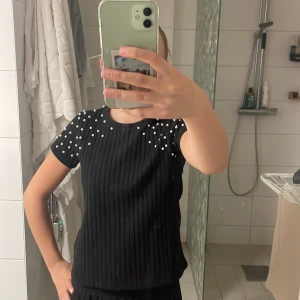 Svart ribbad topp med pärlor - Säljer en svart ribbad topp med korta ärmar och snygga pärldetaljer över axlarna. Toppen är croppad och har en cool, modern vibe som passar perfekt till jeans eller kjol. Materialet känns mjukt och stretchigt, vilket gör den bekväm att bära hela dagen.