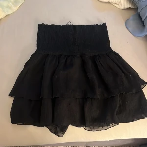 Svart volangkjol med smock - Supersöt svart kortkjol med bred smock i midjan och flera lager volanger. Kjolen har en luftig känsla och är perfekt för dig som gillar en feminin och trendig stil. Materialet känns lätt och flowy. Väldigt stretchig så funkar för flera storlekar 