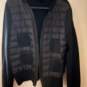 Svart moncler pufferjacka  - Svart pufferjacka från Moncler med quiltad framsida och stickade ärmar. Jackan har fyra framfickor i stickat material, ribbad krage och Moncler-logga på ena ärmen. Dragkedja framtill och stilren, slim passform. Perfekt för höst och vinter.