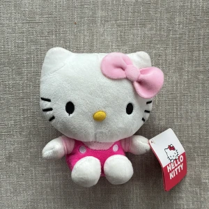 Hello Kitty Plush - Söt Hello Kitty-mjukisdocka i mycket fint skick, med originaletikett kvar. Perfekt för samlare eller som present. Mjuk och ren, inga synliga defekter eller slitage. Klädd i rosa klänning med rosett. En charmig och populär karaktär från Sanrio.