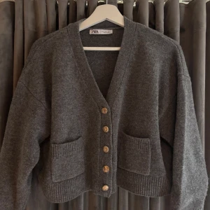 Grå stickad kofta från Zara - Mysig grå stickad kofta från Zara med snygga guldknappar framtill och två stora fickor. Koftan har v-ringning, lång ärm och en loose passform som ger en chill vibe. Perfekt att slänga på sig över en t-shirt eller linne.