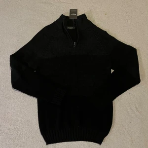 Stickad half zip-tröja - Svart och grå stickad tröja från Dressmann med half zip och hög krage. Tröjan har långärmad design och ribbade muddar vid ärmslut och nederkant. Perfekt för dig som gillar stilren och enkel look.