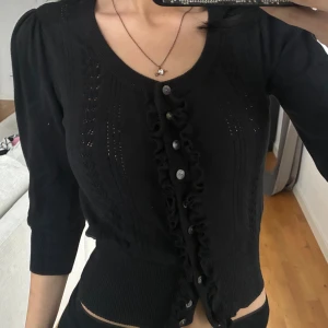 Svart kofta med volang och knappar - Supersöt svart kofta med volangdetaljer längs knappraden och fina stickade mönster framtill. Modellen har trekvartsärm och bred ribbad mudd i midjan för en snygg passform. Perfekt att slänga över en topp för en chic look.