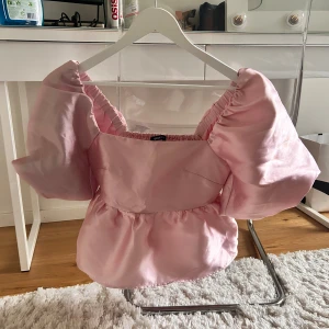 Rosa blus med puffärmar från Gina Tricot - Supersöt ljusrosa blus från Gina Tricot med stora puffärmar och smock i ryggen. Blusen har en fyrkantig urringning och är tillverkad i ett glansigt, satinliknande material. Perfekt för dig som vill sticka ut med en feminin och trendig look.