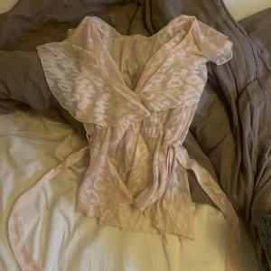 Ljusrosa omlottklänning med mönster - Säljer en söt ljusrosa topp med omlott-design och knytband i midjan. Toppen har ett subtilt leopardmönster och är tillverkad i ett tunt, luftigt material som ger en flowy känsla. Perfekt för dig som gillar romantiska och trendiga detaljer.