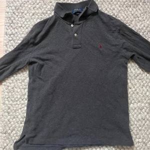 Mörkgrå pikétröja från Ralph Lauren🤩🙌 - Snygg mörkgrå pikétröja från Ralph Lauren med klassisk krage och två knappar framtill. Tröjan har lång ärm och den ikoniska röda Polo-loggan broderad på bröstet. Passar S/xs