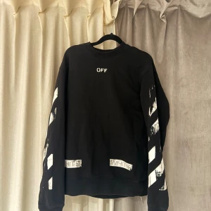 Svart Off-White sweatshirt med tryck - Svart sweatshirt från Off-White med vita, slitna ränder och texttryck på både ärmar och rygg. Tröjan har en klassisk rund hals och lång ärm, tillverkad i mjuk bomull. Snygg streetwear-vibe med tydliga Off-White detaljer och grafiskt mönster.