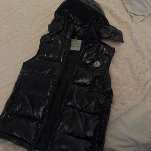 Svart dunväst från Moncler med huva - Snygg svart glansig dunväst från Moncler med avtagbar huva. Västen har dragkedja framtill, två fickor med dragkedja och moncler logga. Otroligt skön och i nyskick!! 🖤 Fraktar snabbt! Jag har kunnat haft den på vintern också med varmare tröjor under